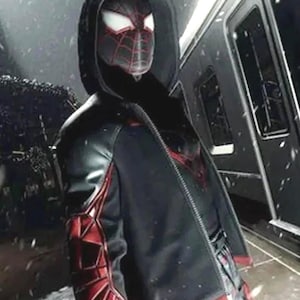 May include: A black hooded jacket with red and black accents, featuring a spider-themed design. The jacket has a zipper closure and is worn over a patterned shirt. The image is set against a snowy background.