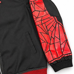 May include: Black jacket with a red geometric design on the right sleeve. The jacket has a black collar and a red ribbed hem. The sleeve design features red shapes outlined in black. The jacket has a zipper closure and a pocket.