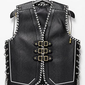 May include: A black waistcoat with a snakeskin-like texture, featuring three gold-toned buckles across the front, white and black braided trim along the edges, and side lacing with matching braided cords and gold-toned metal aglets at the ends.