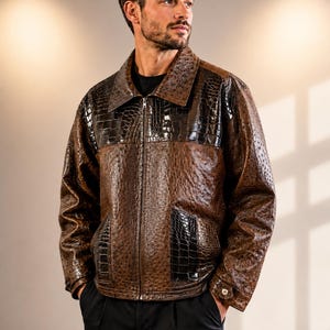 Può includere: Una giacca marrone caratterizzata da una texture effetto struzzo sul corpo e sulle maniche, a contrasto con pannelli in pelle marrone scuro con stampa coccodrillo sulla parte superiore del petto e sulle tasche. La giacca ha una zip frontale.