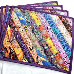 May include: A set of colorful, quilted placemats with a vibrant, abstract design. Each placemat features a patchwork of patterned fabric strips in shades of orange, yellow, blue, and purple, bordered with a solid purple trim. The placemats are arranged in a fan shape.