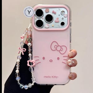 May include: Pink phone case featuring a Hello Kitty design. The case has a clear border and a decorative strap with beads and charms. The Hello Kitty face and bow are visible on the case, with the text "-Hello Kitty-" below.