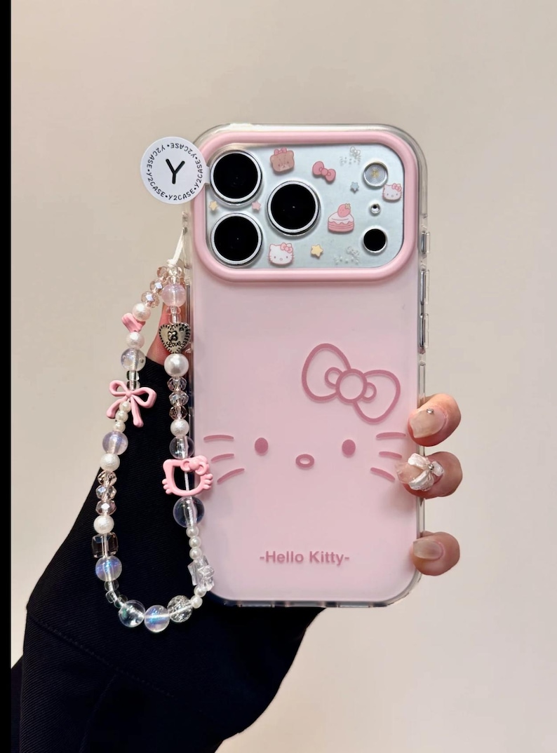 May include: Pink phone case featuring a Hello Kitty design. The case has a clear border and a decorative strap with beads and charms. The Hello Kitty face and bow are visible on the case, with the text "-Hello Kitty-" below.