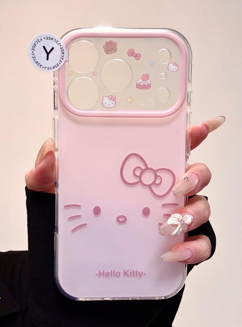 May include: Pink phone case featuring a Hello Kitty design. The case has a clear border and a pink back with a Hello Kitty face, bow, and other decorative elements. The text "-Hello Kitty-" is printed at the bottom.