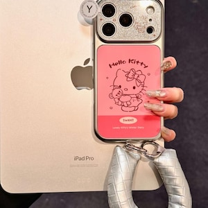 Op de afbeelding: Roze en zilveren telefoonhoesje met een Hello Kitty-ontwerp. De hoes heeft een roze paneel met een Hello Kitty-afbeelding en de tekst "Hello Kitty". Een zilveren gevlochten polsband is bevestigd.
