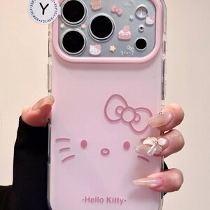 May include: Pink phone case featuring a Hello Kitty design. The case has a clear border and a pink back with a Hello Kitty face and bow. The top section has decorative elements including a cake and stars. The text "-Hello Kitty-" is printed at the bottom.