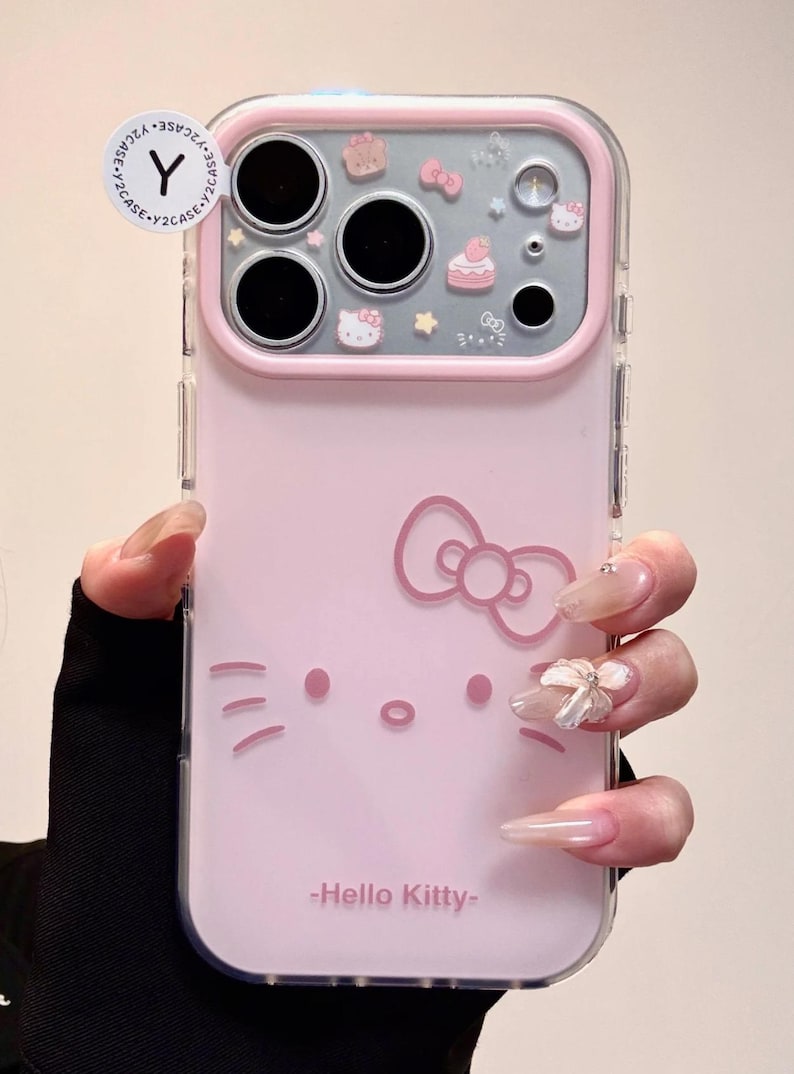 May include: Pink phone case featuring a Hello Kitty design. The case has a clear border and a pink back with a Hello Kitty face and bow. The top section has decorative elements including a cake and stars. The text "-Hello Kitty-" is printed at the bottom.