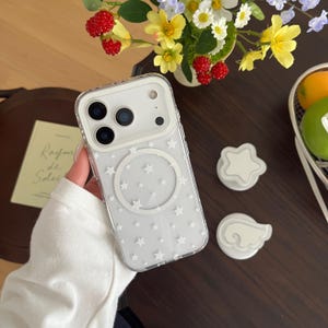 以下が含まれることがあります： 白いMagSafeリングと小さな白い星が散りばめられた模様が特徴の透明なスマートフォンケース。ケースには白いカメラモジュール枠があり、3つの円形のレンズ用切り抜きとフラッシュ用の開口部が備わっています。