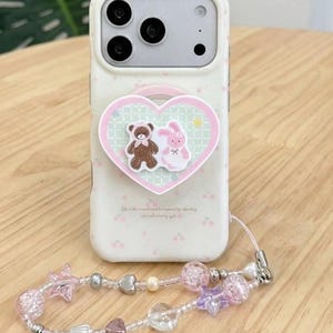 May include: A white smartphone case with a faint cherry pattern and a heart-shaped pop-out grip featuring a brown bear and a pink rabbit. Attached is a beaded phone charm with pink and purple star-shaped beads, clear spheres, and small metallic hearts.