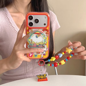 May include: A yellow smartphone case featuring Toy Story character graphics, a clear circular shaker charm with green alien figures, and a colourful beaded wrist strap with a dangling red plastic charm that reads Toy Story in blue and yellow letters.