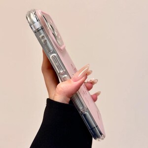 May include: A clear phone case with a pink interior and a silver border. The case is held at an angle, showcasing its design. The phone case is a protective accessory for smartphones.