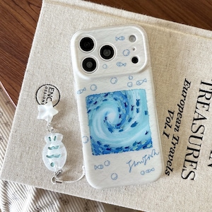 May include: A white smartphone case featuring a central square illustration of a blue swirling ocean with small fish. The case is decorated with tiny blue fish and bubble icons, and includes a dangling charm with a white star and a fish shape.