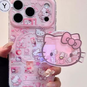 以下が含まれることがあります： ピンク色のハローキティデザインの携帯電話ケース。ケースには複数のハローキティのイラストと透明な保護カバーが付いています。お揃いのハローキティの携帯電話グリップも表示されています。
