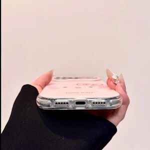 May include: A pink phone case with a clear border. The case features a Hello Kitty design with the text "Hello Kitty." The case is designed to fit a smartphone and has cutouts for the charging port and speakers.