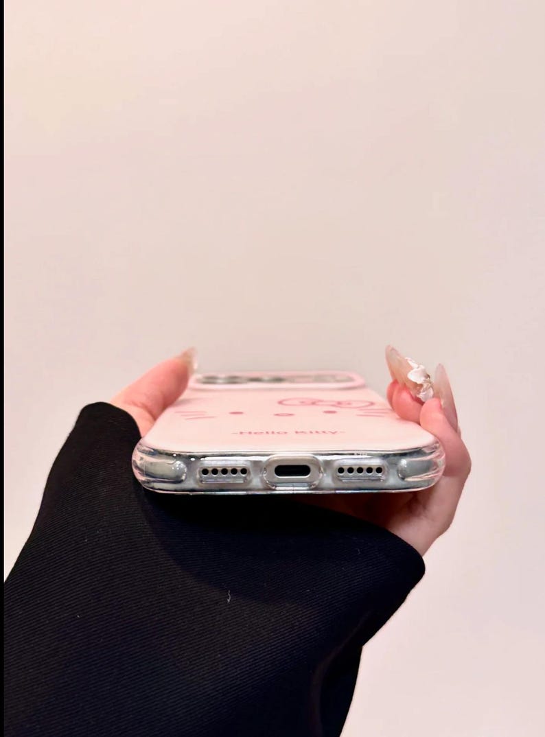 May include: A pink phone case with a clear border. The case features a Hello Kitty design with the text "Hello Kitty." The case is designed to fit a smartphone and has cutouts for the charging port and speakers.