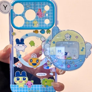 May include: A transparent phone case featuring a blue and white checked design and cartoon characters. A matching charm in the shape of a retro digital pet game is attached. The text "たまごっち" is visible.