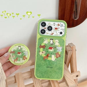 May include: A light green smartphone case and a matching circular phone grip. Both items feature a printed pattern of various cartoon animals, including dogs, cats, a rabbit, and an elephant, arranged around a small red-roofed house illustration.