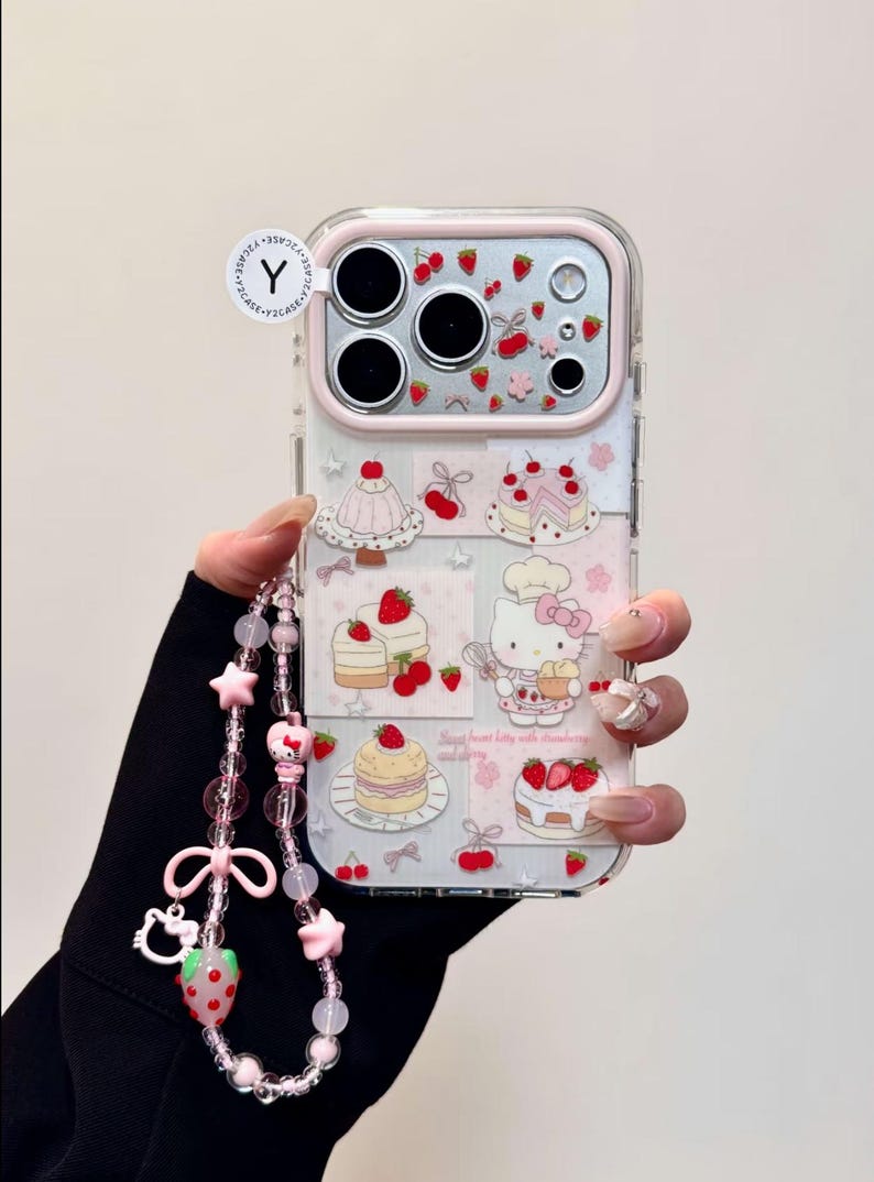 May include: Clear phone case with a pink border featuring a collage of strawberry and cake illustrations. A pink and clear beaded wrist strap with star and bow charms is attached. The case has a Hello Kitty theme.