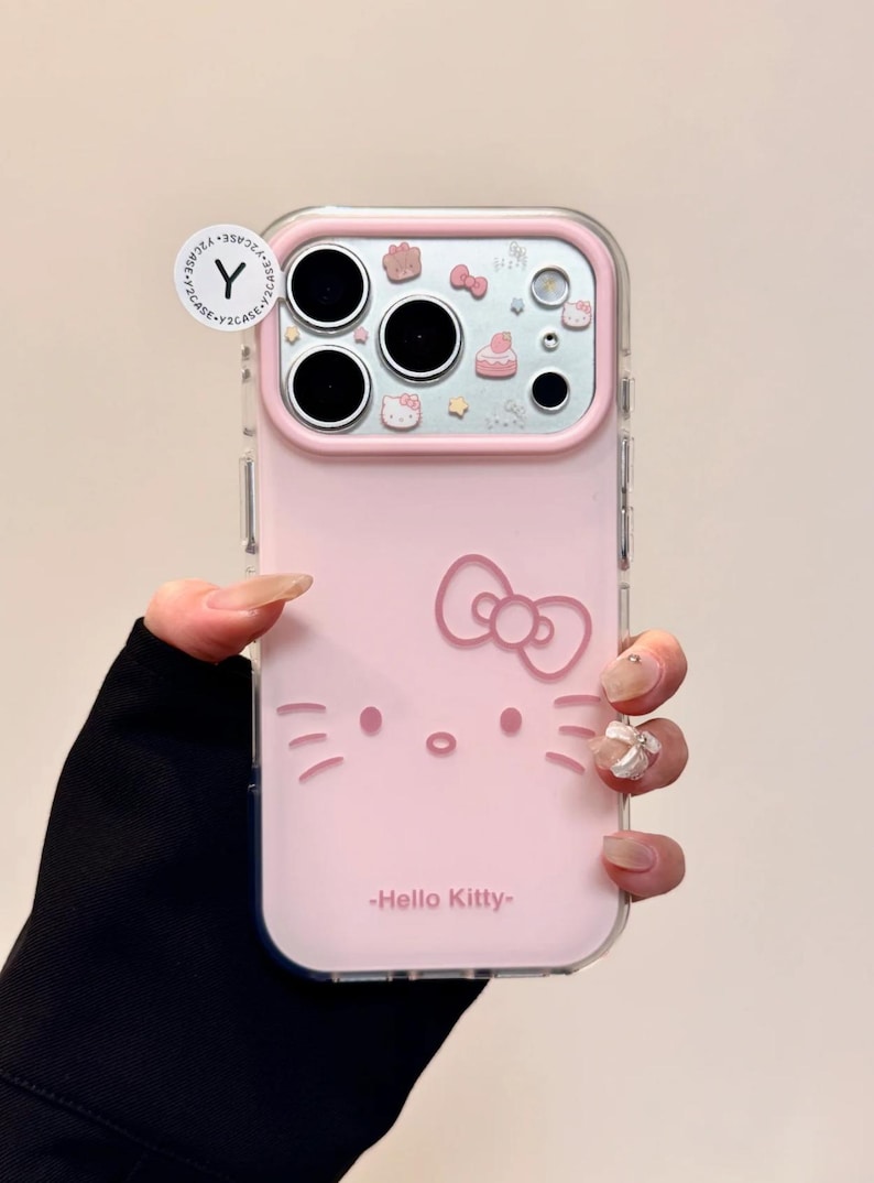 May include: Pink phone case featuring a Hello Kitty design. The case has a clear border and includes a bow, face, and the text "Hello Kitty". The camera area has decorative elements.