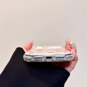 May include: Clear phone case with a pattern of small red hearts. The case is held in a hand, showing the bottom edge with speaker and charging ports. The case is designed to fit a smartphone.