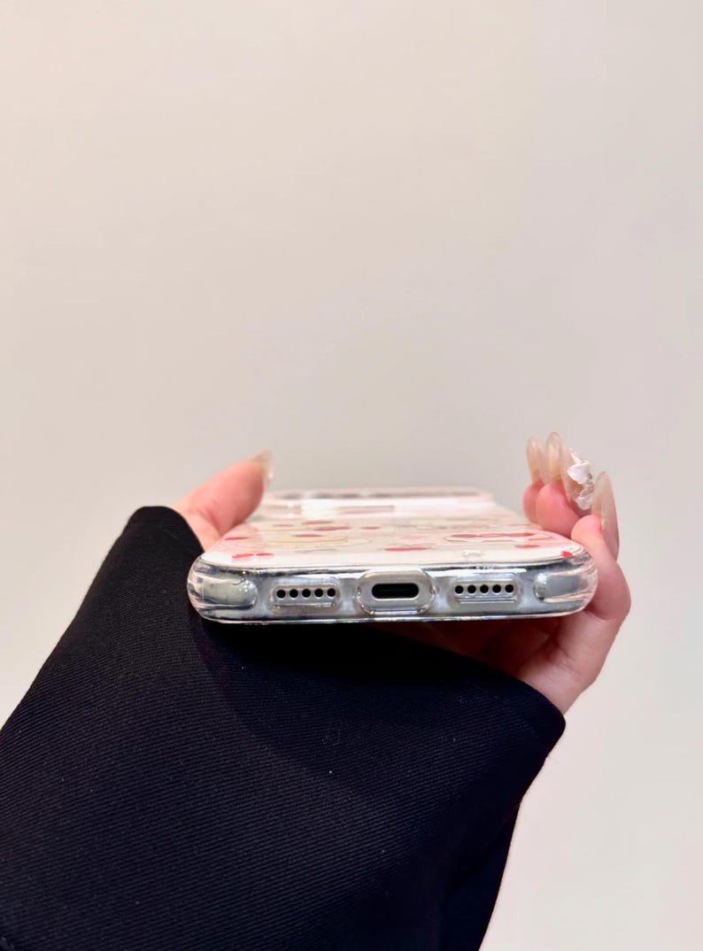 May include: Clear phone case with a pattern of small red hearts. The case is held in a hand, showing the bottom edge with speaker and charging ports. The case is designed to fit a smartphone.