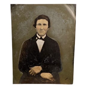 May include: A vintage tintype photograph featuring a portrait of a person with dark hair, wearing a dark coat over a white collared shirt with a black bow tie. The image has a weathered, metallic texture with visible surface wear and discoloration.