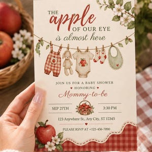 以下が含まれることがあります： THE apple OF OUR EYE is almost hereというテキストが記載された長方形のベビーシャワー招待状。物干しロープにかかったベビー服、リンゴのバスケット、下部の赤いギンガムチェックの縁取りのイラストが描かれています。
