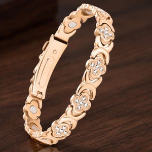 May include: A gold-colored bracelet with a floral design. The bracelet features multiple flower-shaped accents, each adorned with small, clear crystals. The clasp is a simple, rectangular design. The bracelet is displayed on a dark wooden surface.