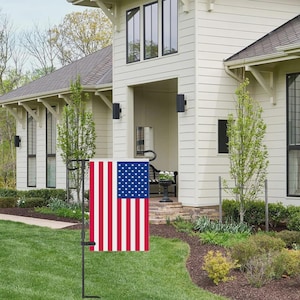 May include: A rectangular garden flag featuring the United States flag design with red and white vertical stripes and a blue canton with white stars, suspended from a black metal garden flag stand positioned in a green lawn.