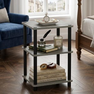 May include: A three-tier side table with a gray wood-grain finish and black cylindrical supports. The top shelf holds a small clock and a decorative dish. The middle shelf features a book and a candle. The bottom shelf holds two stacked books.