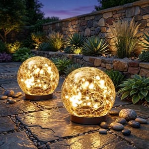 May include: Two spherical outdoor lights with a crackled glass texture and warm internal illumination. Each globe sits on a dark circular base, casting a soft glow on the surrounding stone patio surface and nearby garden plants.