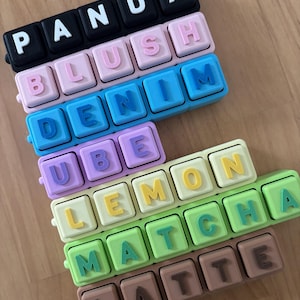 以下が含まれることがあります： 「PANDA」、「BLUSH」、「DENIM」、「UBE」、「LEMON」、「MATCHA」、「LATTE」などの単語が書かれた、カラフルなプラスチック製のワードタイル。タイルは様々なパステルカラーで、白と黒の文字が使われています。