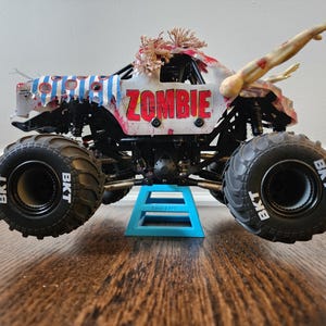 May include: A toy monster truck with large black tyres and the word "ZOMBIE" in red letters. The truck has a white body with blue and red accents, and a zombie arm extending from the top.