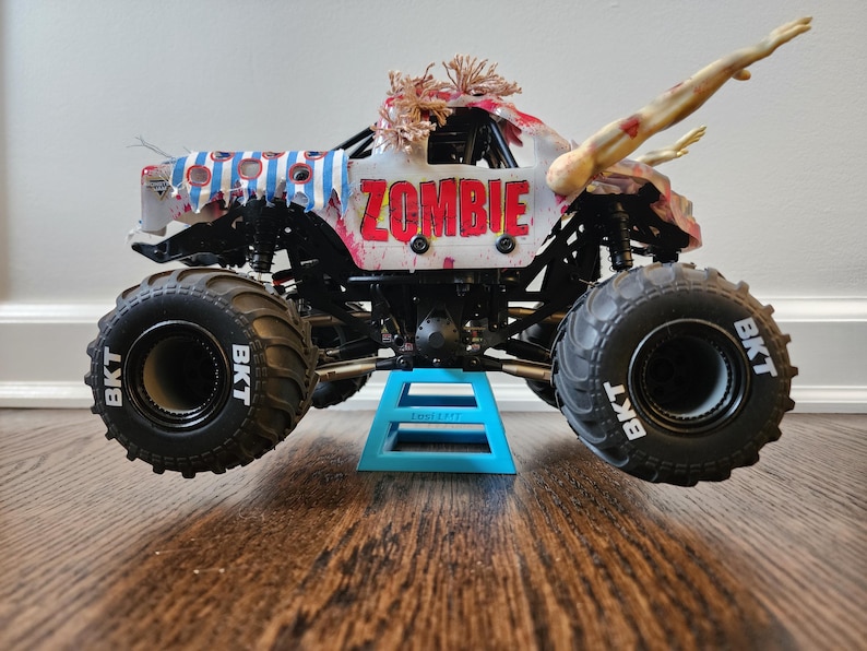 May include: A toy monster truck with large black tyres and the word "ZOMBIE" in red letters. The truck has a white body with blue and red accents, and a zombie arm extending from the top.
