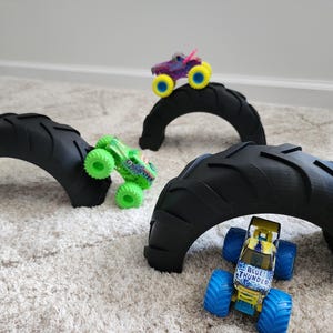 以下が含まれることがあります： おもちゃのモンスタートラックが置かれた、黒いタイヤアーチが3つ。緑色のモンスタートラックが左に、紫と黄色のトラックが中央のアーチの上に、青と黄色のトラックには「Blue Thunder」の文字が書かれており、右のアーチの下にあります。