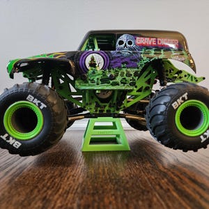 May include: A green and black Grave Digger monster truck toy with large black tyres and green rims. The truck has a skull graphic and the words "Grave Digger" on the roof. The truck is on a green stand.