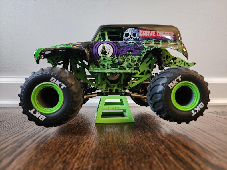 May include: A green and black Grave Digger monster truck toy with large black tyres and green rims. The truck has a skull graphic and the words "Grave Digger" on the roof. The truck is on a green stand.
