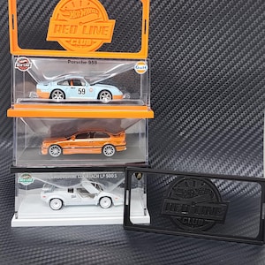 Puede incluir: Tres modelos de coches Hot Wheels Red Line Club en vitrinas. Los coches son un Porsche 959 azul claro, un coche naranja y un Lamborghini Countach LP 500 S blanco. Las vitrinas están apiladas en un marco naranja.