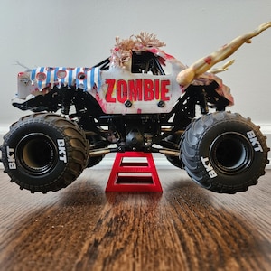 May include: A toy monster truck, white with blue stripes and the word "ZOMBIE" in red. It has large black tyres with "BKT" on them. The truck is supported by a red stand and features a zombie arm.