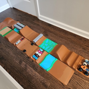 Puede incluir: Un conjunto de pista de juguete de plástico marrón con múltiples rampas, colinas y secciones texturizadas en verde y azul. Varios coches de juguete pequeños y dos camiones monstruo con ruedas grandes están colocados a lo largo de la pista.