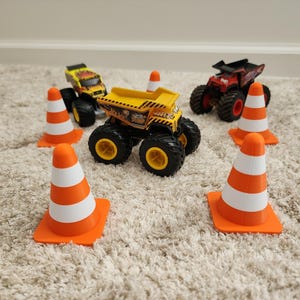 May include: A collection of toy monster trucks and orange and white traffic cones. The trucks are yellow, red, and black with large tires. The cones are arranged around the trucks, suggesting a construction or obstacle course theme.