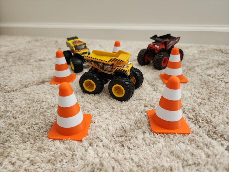 May include: A collection of toy monster trucks and orange and white traffic cones. The trucks are yellow, red, and black with large tires. The cones are arranged around the trucks, suggesting a construction or obstacle course theme.