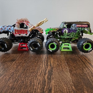 May include: Two toy monster trucks. One is labelled "ZOMBIE" with a zombie design, the other "GRAVE DIGGER" with a skull graphic. Both have large black tyres and are on small stands.