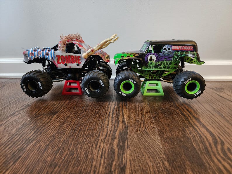 May include: Two toy monster trucks. One is labelled "ZOMBIE" with a zombie design, the other "GRAVE DIGGER" with a skull graphic. Both have large black tyres and are on small stands.