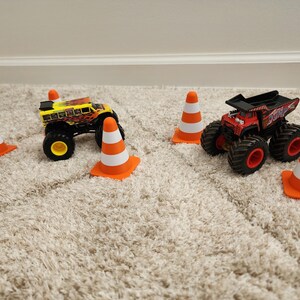 May include: Two toy monster trucks, one yellow and one red, are surrounded by orange and white traffic cones. The trucks have large black tires and are positioned on a beige carpet.