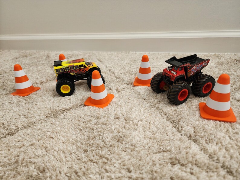 May include: Two toy monster trucks, one yellow and one red, are surrounded by orange and white traffic cones. The trucks have large black tires and are positioned on a beige carpet.
