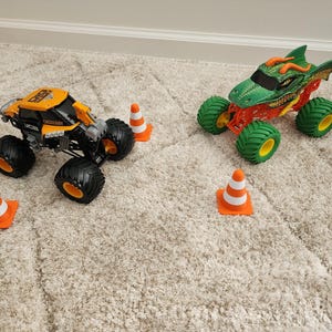 May include: Two toy monster trucks, one orange and black, the other green and red, sit on a carpet. The orange truck has black tires and orange rims. The green truck is designed to look like a dragon. Orange and white traffic cones are scattered around the trucks.