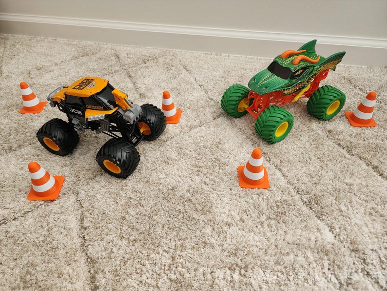 May include: Two toy monster trucks, one orange and black, the other green and red, sit on a carpet. The orange truck has black tires and orange rims. The green truck is designed to look like a dragon. Orange and white traffic cones are scattered around the trucks.