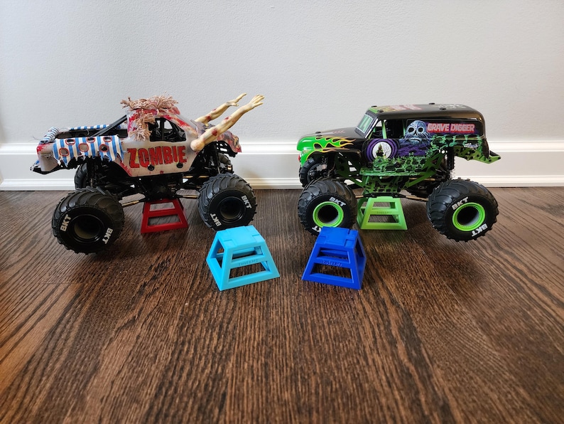 May include: Two toy monster trucks, one labelled "ZOMBIE" with a zombie design and the other "GRAVE DIGGER" with a skull graphic. Both trucks are on stands, with two blue stands in front.