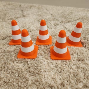 May include: Five orange and white striped traffic cones. Each cone has a square orange base and a conical top with three white stripes. The cones are arranged on a beige carpet.